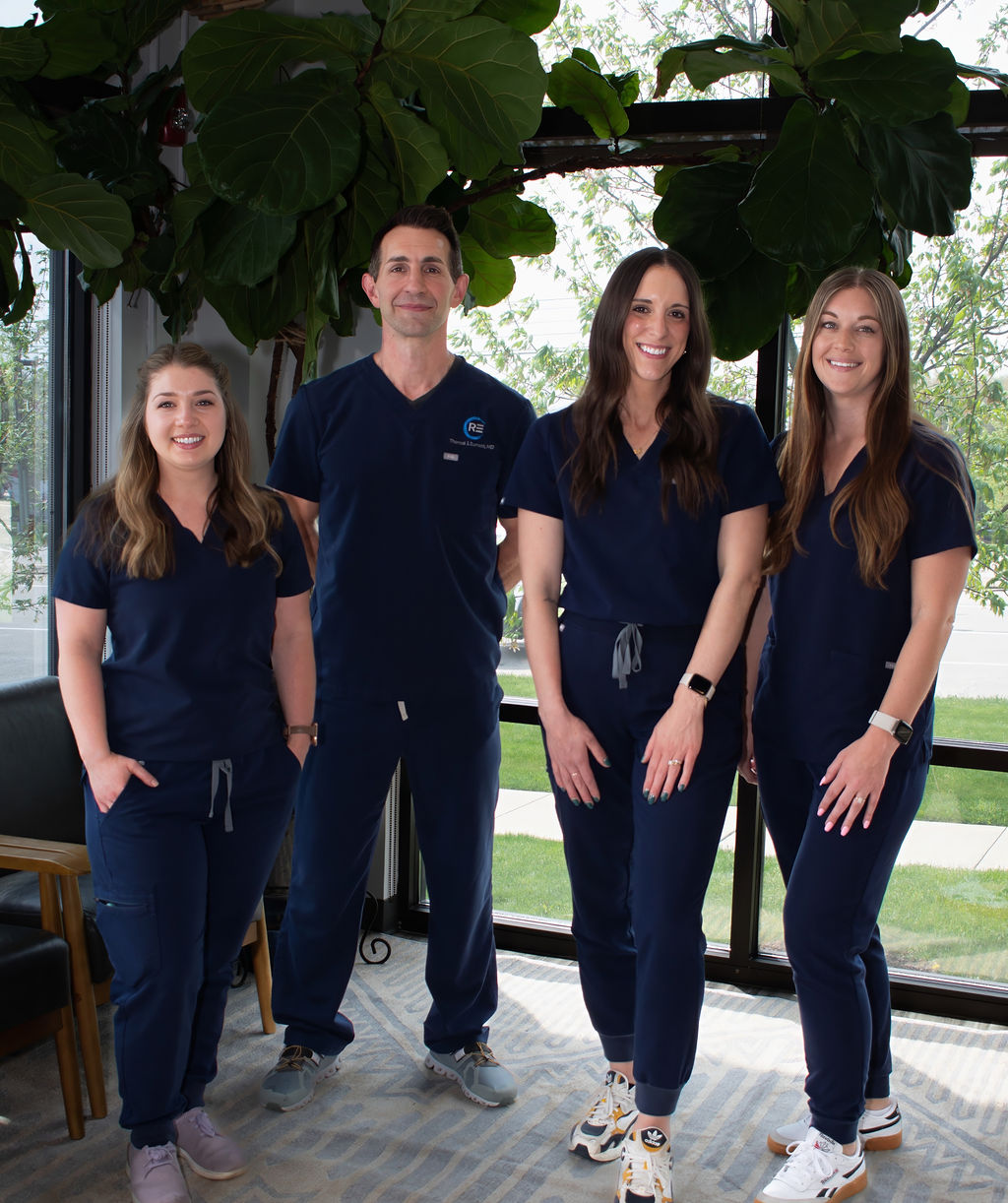 Group of four people in matching navy blue scrubs standing indoors with large windows and greenery.