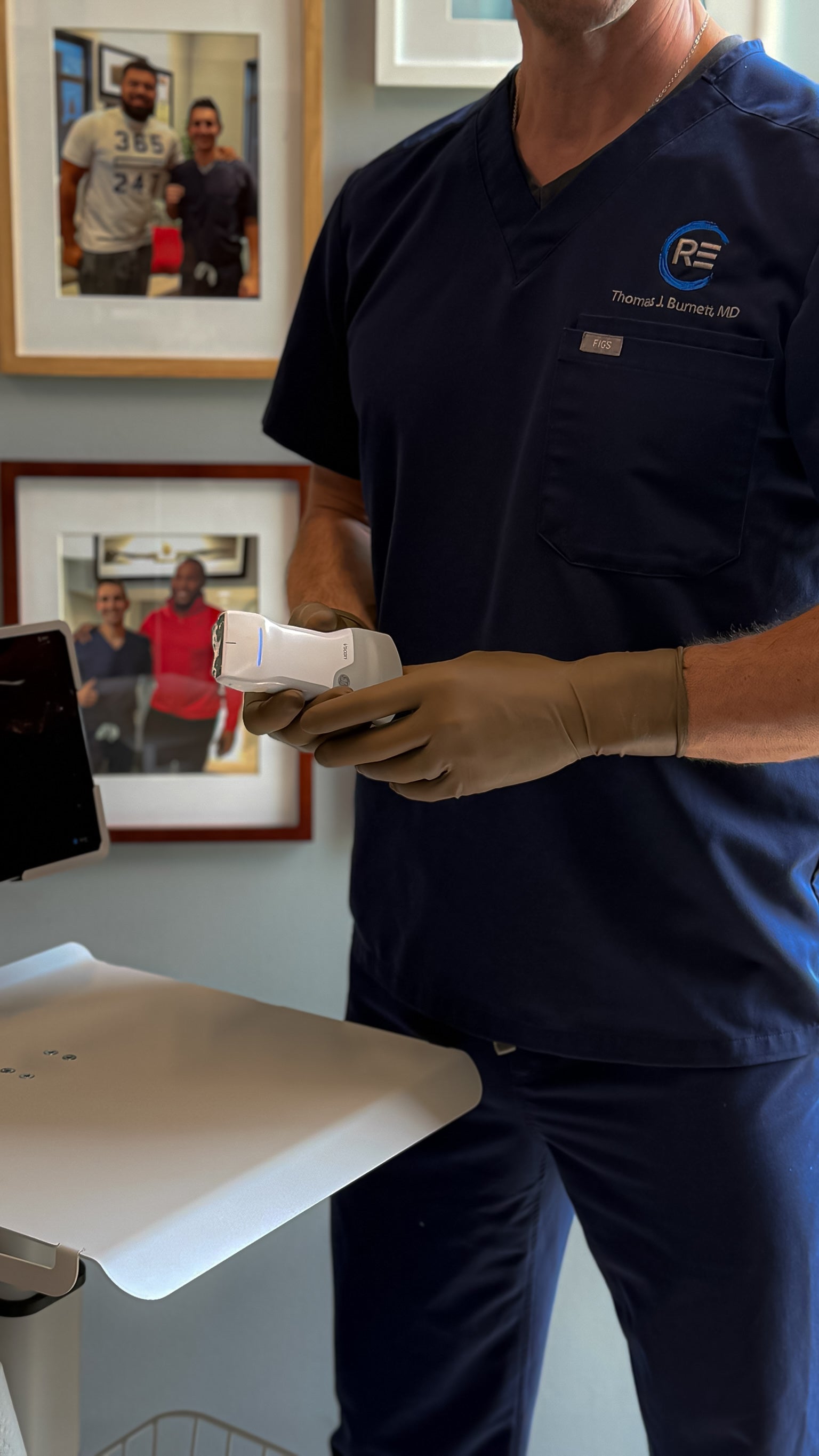 Person in medical scrubs holding a device in a clinical setting with framed pictures on the wall.