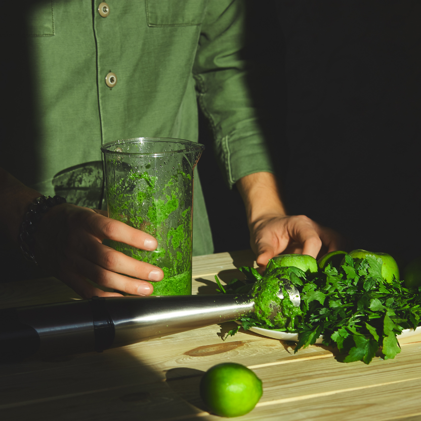Person preparing a green smoothie with a blender on a wooden surface.