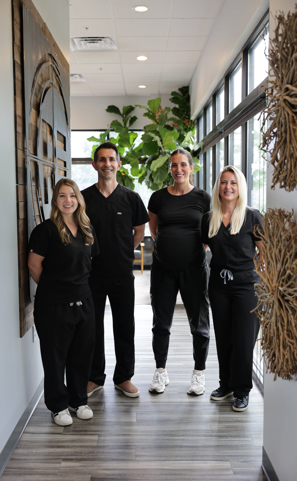 Four people wearing matching black medical scrubs and lounge sets standing together in a brightly lit hallway with large windows and indoor plants.