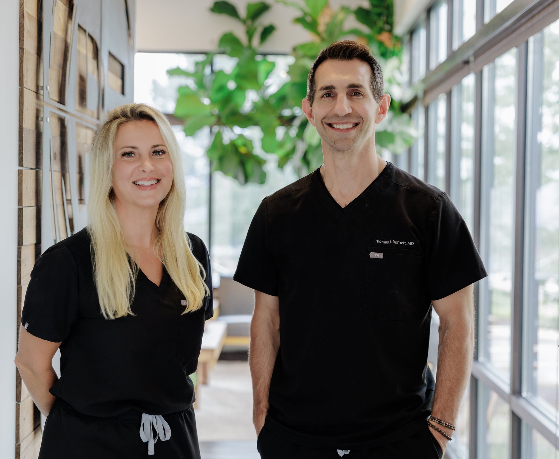 A smiling blonde woman and a smiling man standing side by side in matching black V-neck medical scrubs inside a bright modern office or clinic with large windows and indoor plants.