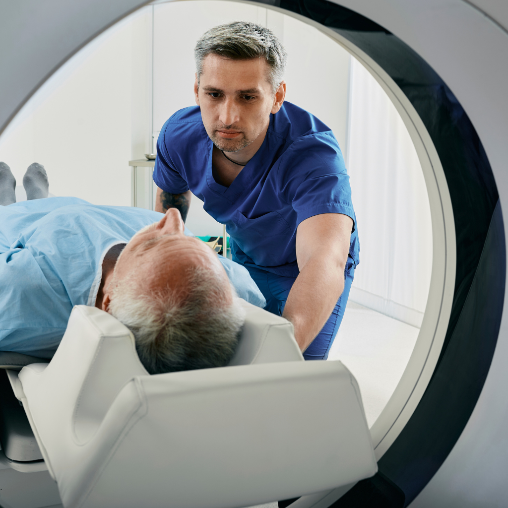 A doctor in blue scrubs leans over a patient lying on a CT scanner table, viewed through the circular opening of the machine in a bright clinical room.