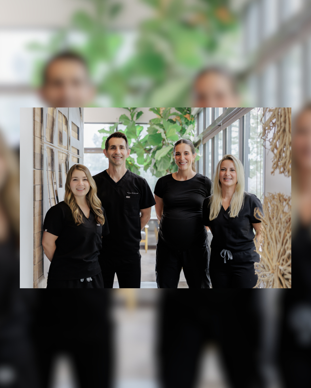 Four people wearing matching black scrubs standing together in an indoor setting with large windows and greenery in the background.