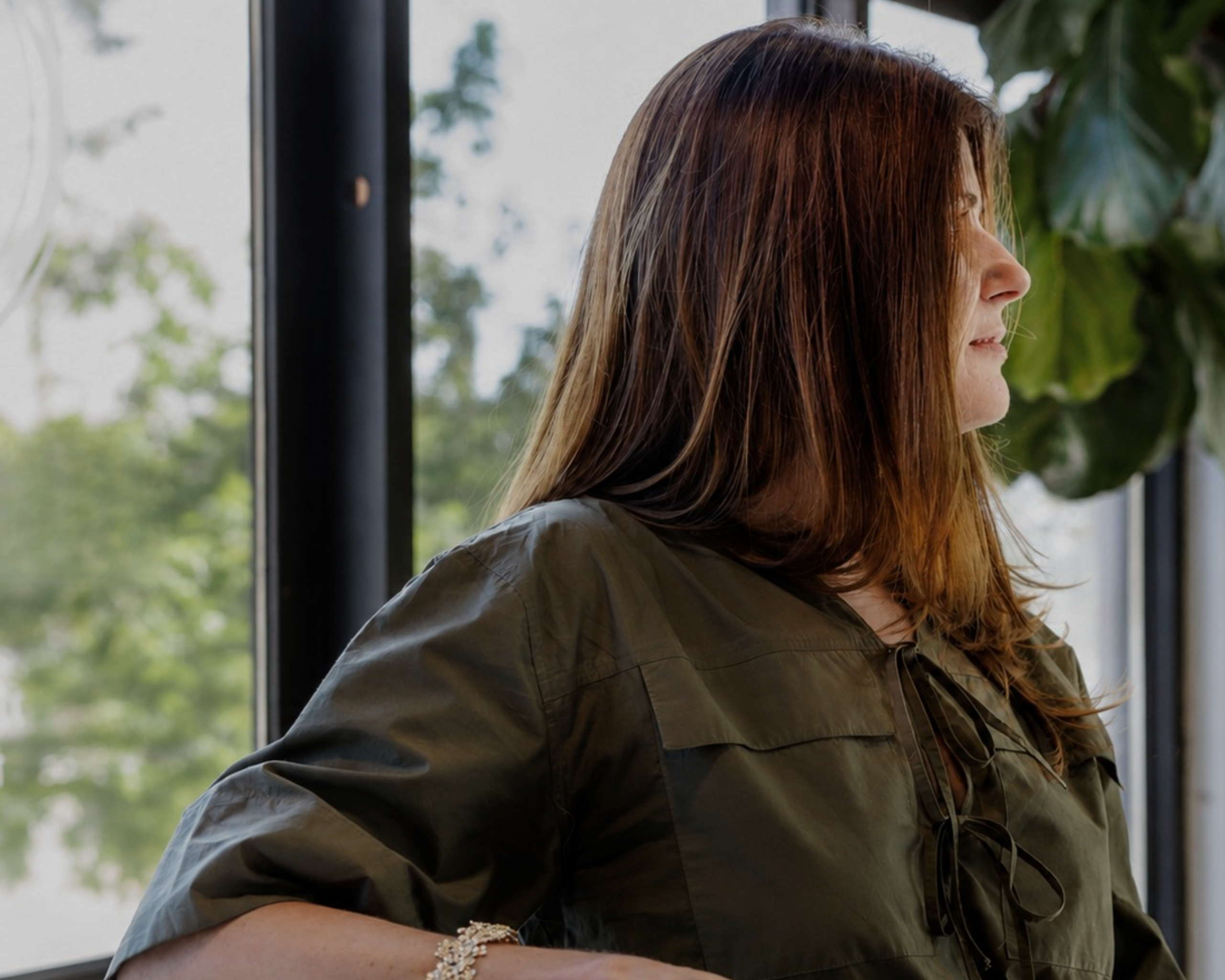 A woman with long brown hair is shown in profile, wearing an olive green blouse with tie details at the neckline, set against a window background with soft natural light.