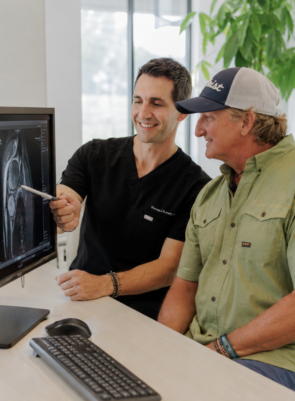 A doctor in black medical scrubs points at an MRI scan on a computer monitor, while a patient wearing an olive green button-up shirt and a baseball cap sits beside him at a desk.