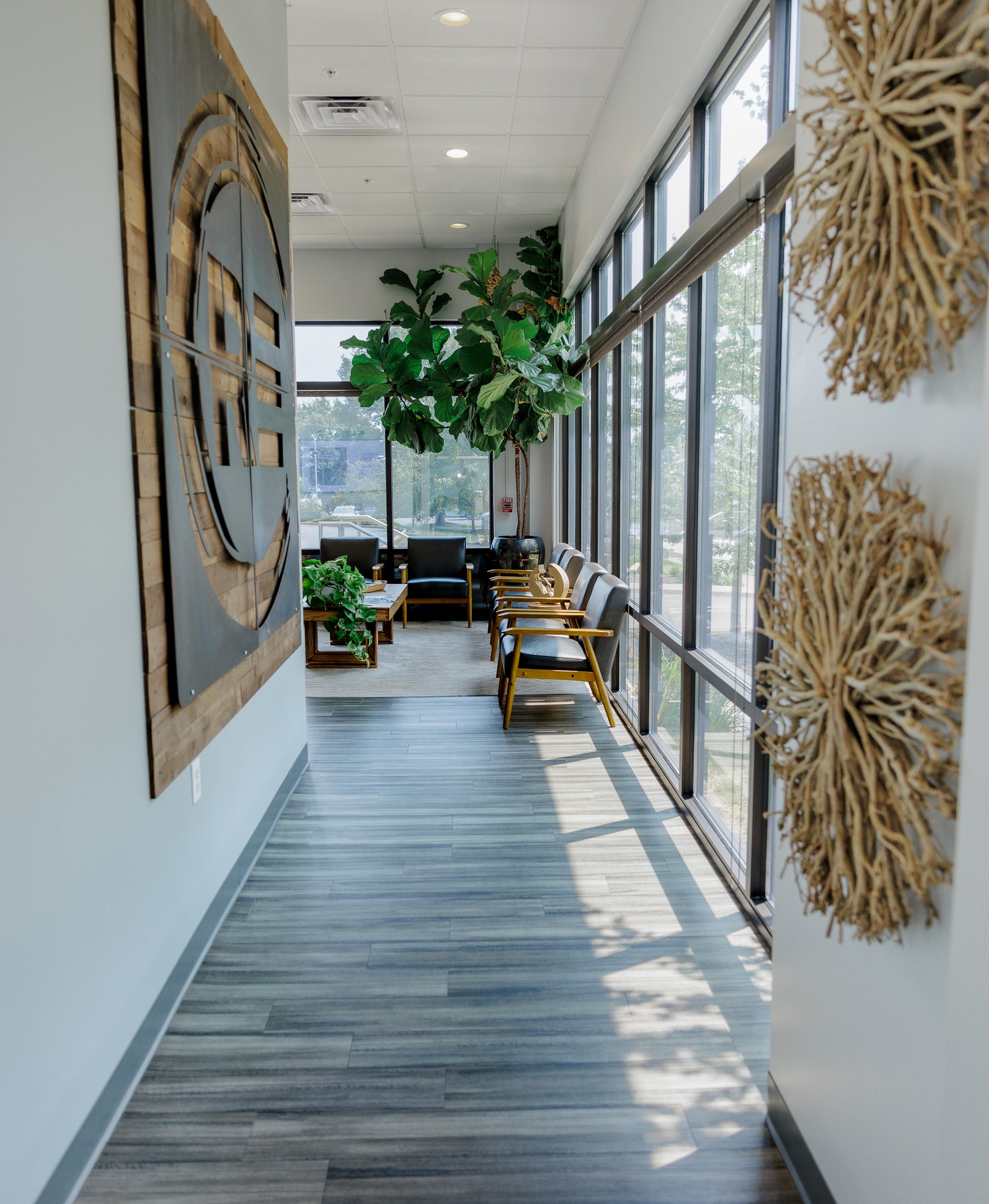 A modern interior hallway with gray wood-look flooring, large windows, lounge chairs, potted plants, and decorative wall art.