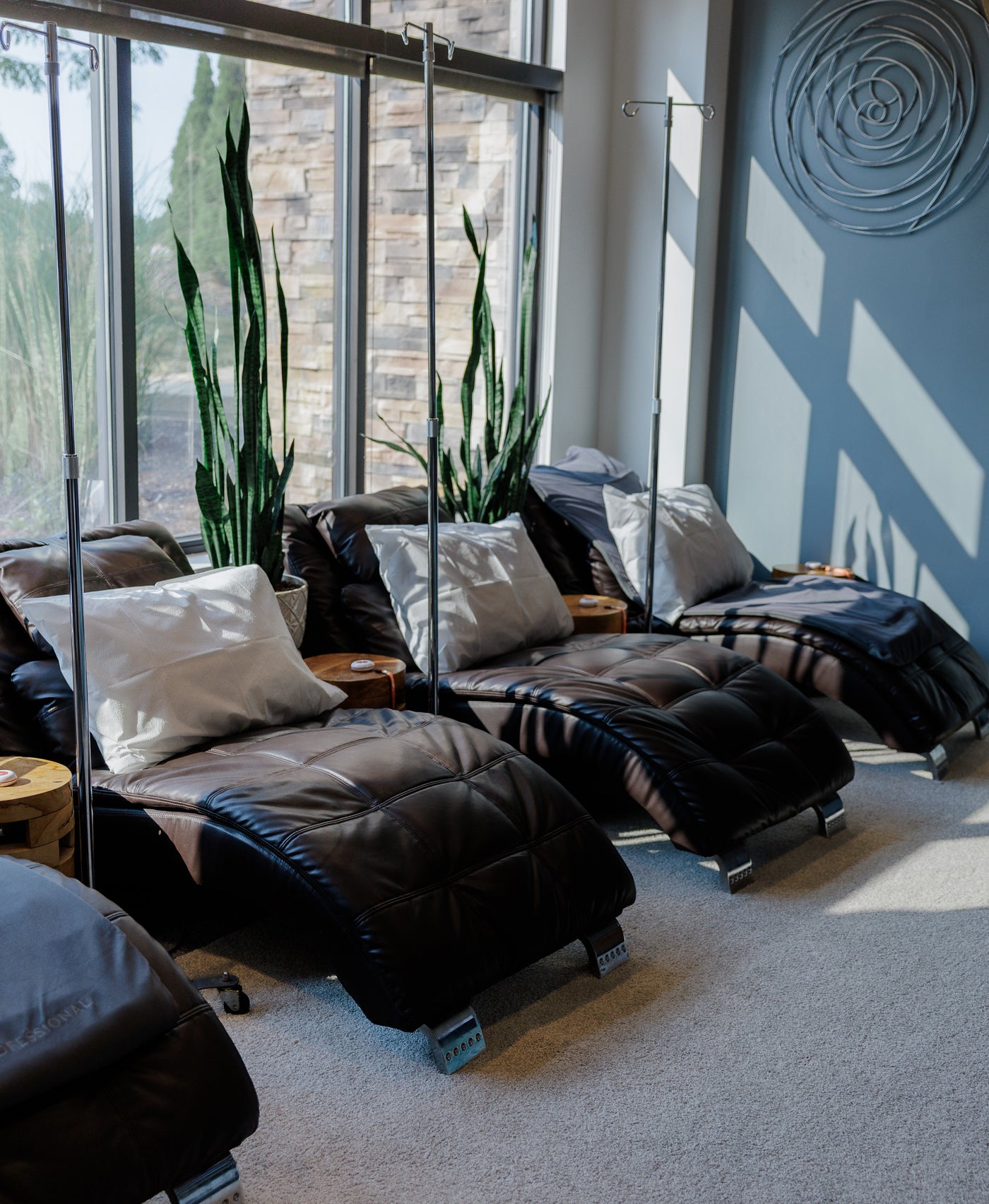 A modern spa-style lounge area with three dark brown leather chaise lounges arranged near large windows, accented with white and gray pillows, with a gray wall and decorative circular wall art in the background.