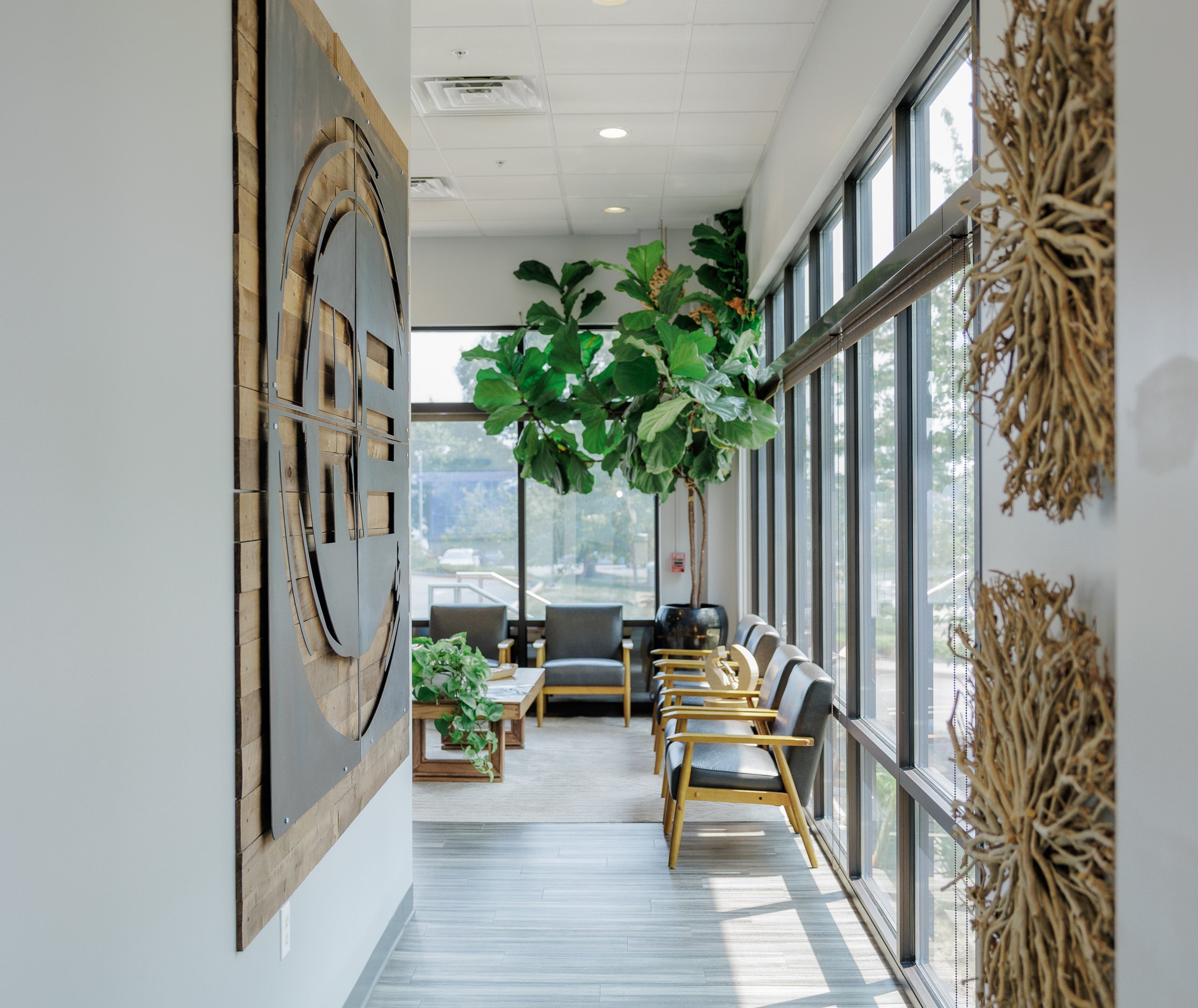 A modern office lobby or lounge area with tall floor-to-ceiling windows, gray upholstered chairs with wood frames, potted green plants, and large decorative wall art mounted on white walls.