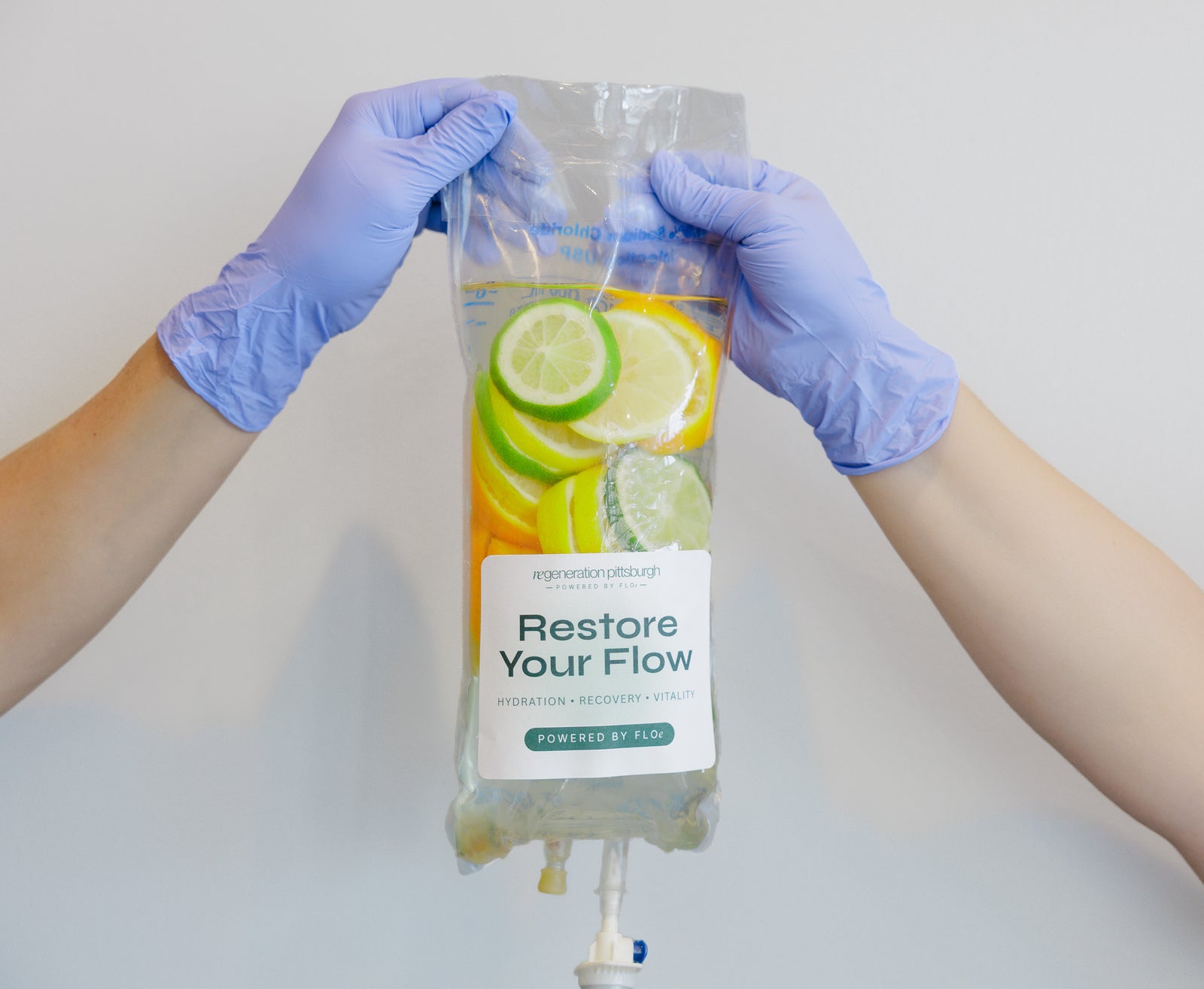 A person wearing blue nitrile gloves holds a clear IV-style hydration bag labeled 'Restore Your Flow' against a plain light gray background. The bag contains pale yellow liquid with visible lime and lemon slices printed on the front.