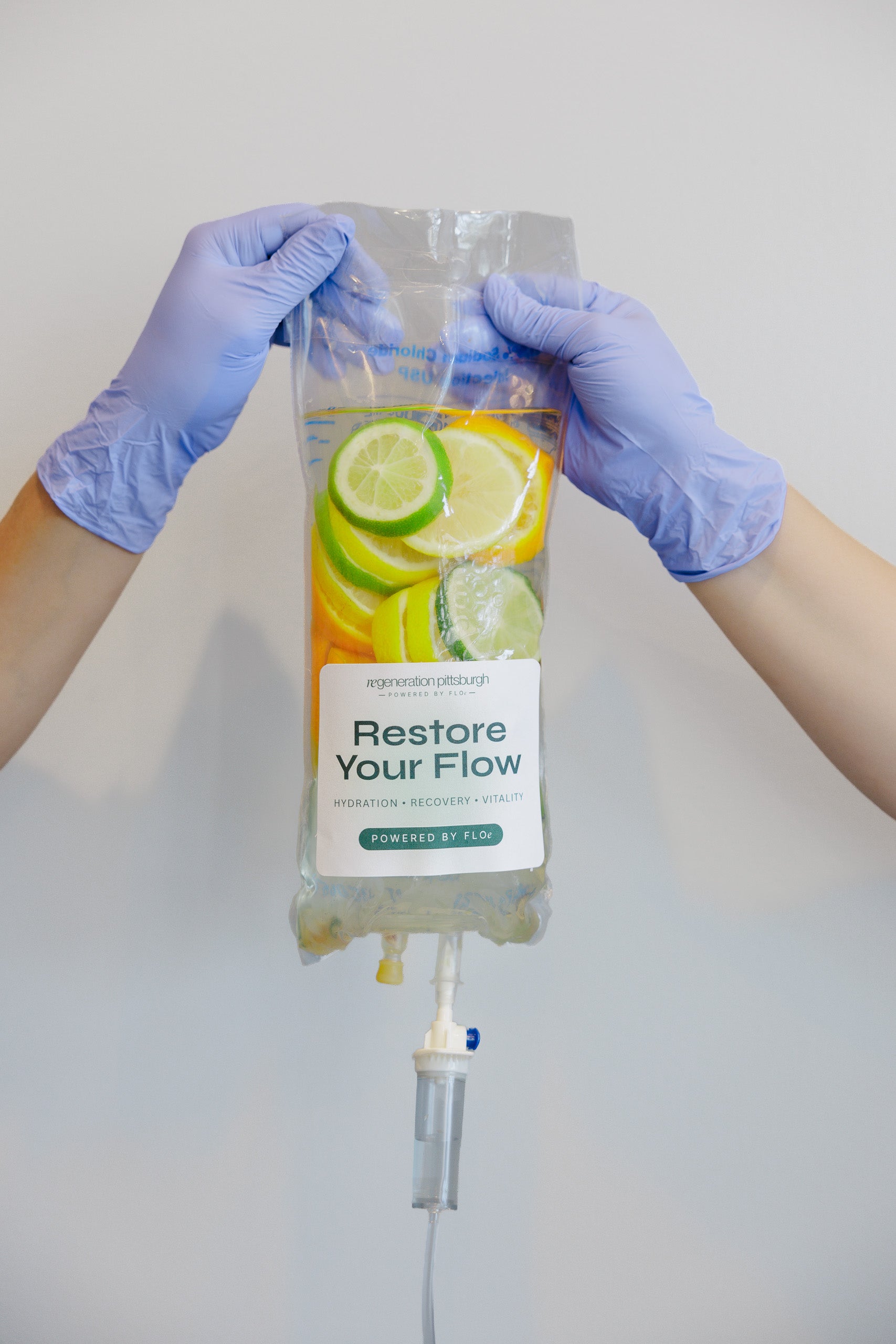 A clear IV-style pouch filled with yellow and green citrus slices, held up by two gloved hands against a plain light gray background. The pouch label reads 'Restore Your Flow' and 'Purified by AIOS'.