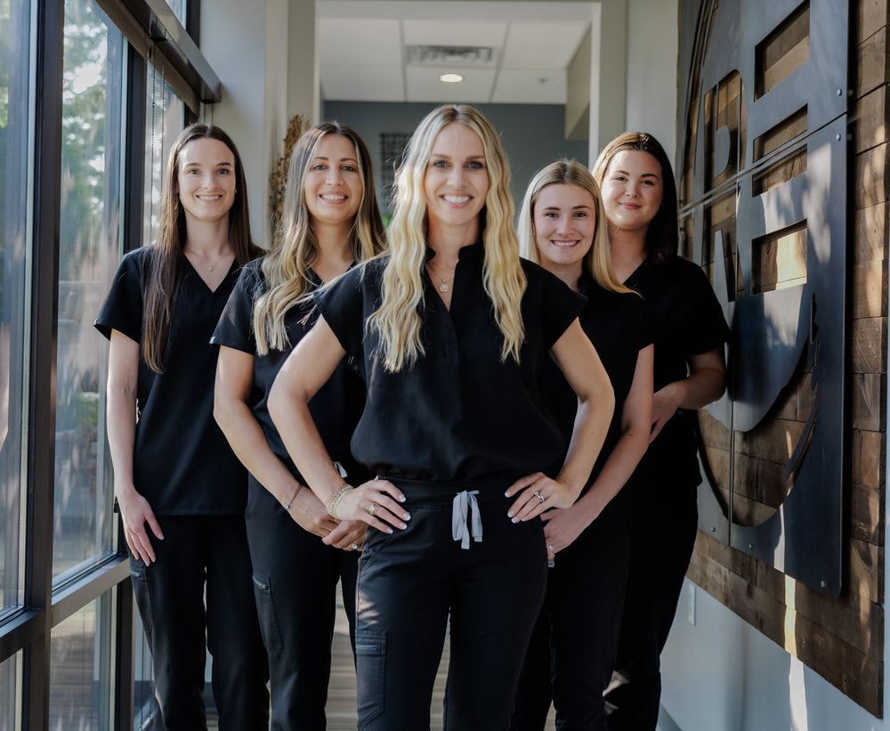 Five women standing together in a hallway, all wearing matching black scrubs or black tops and dark pants. The setting appears to be an indoor office or clinic corridor with large windows on the left and a wood-paneled wall on the right.