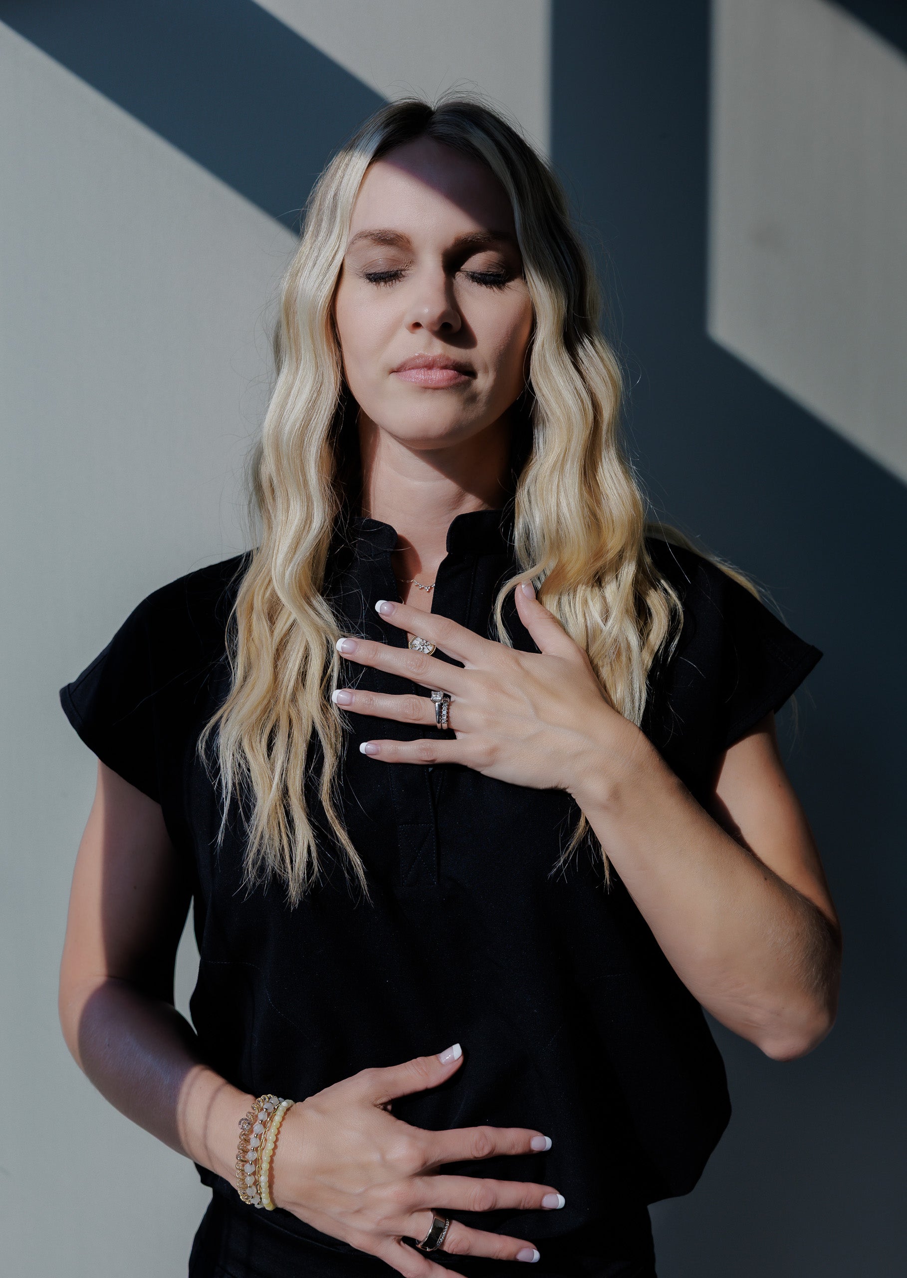 A blonde woman with her eyes closed, wearing a black short-sleeve button-up shirt and gold bracelets, posing with her hands on her chest against a gray wall with diagonal shadows.