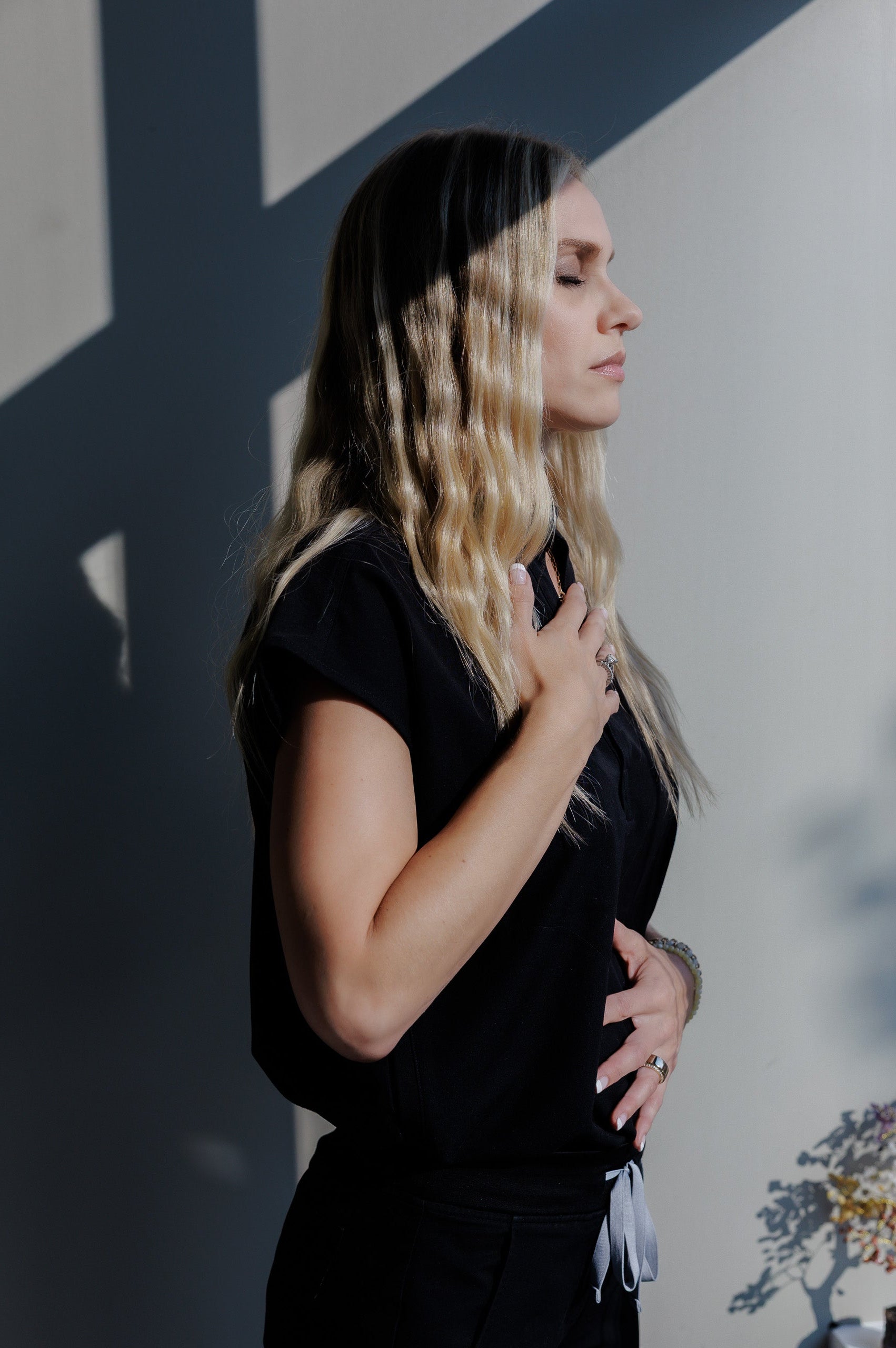 A woman with blonde highlights and dark roots is shown in profile wearing a black short-sleeve top and black bottoms, standing against a gray wall with dramatic shadows.
