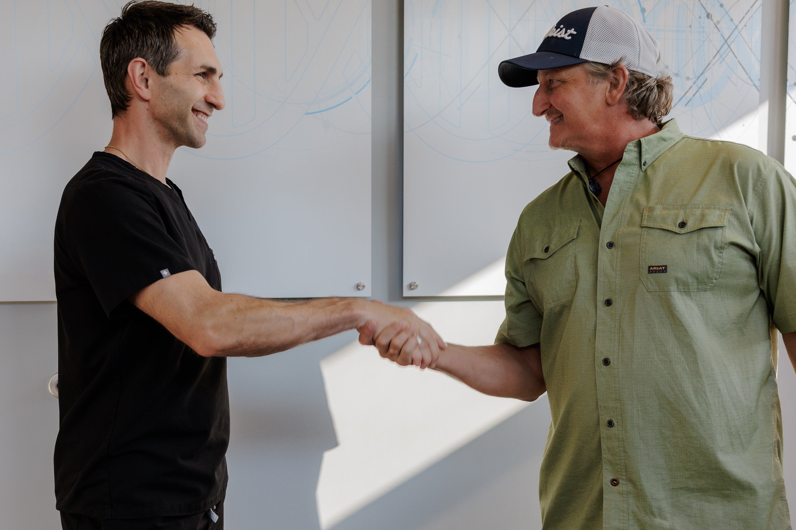 Two men shaking hands in a bright indoor setting. The man on the left wears a black short-sleeve t-shirt, and the man on the right wears an olive green button-up shirt and a baseball cap.
