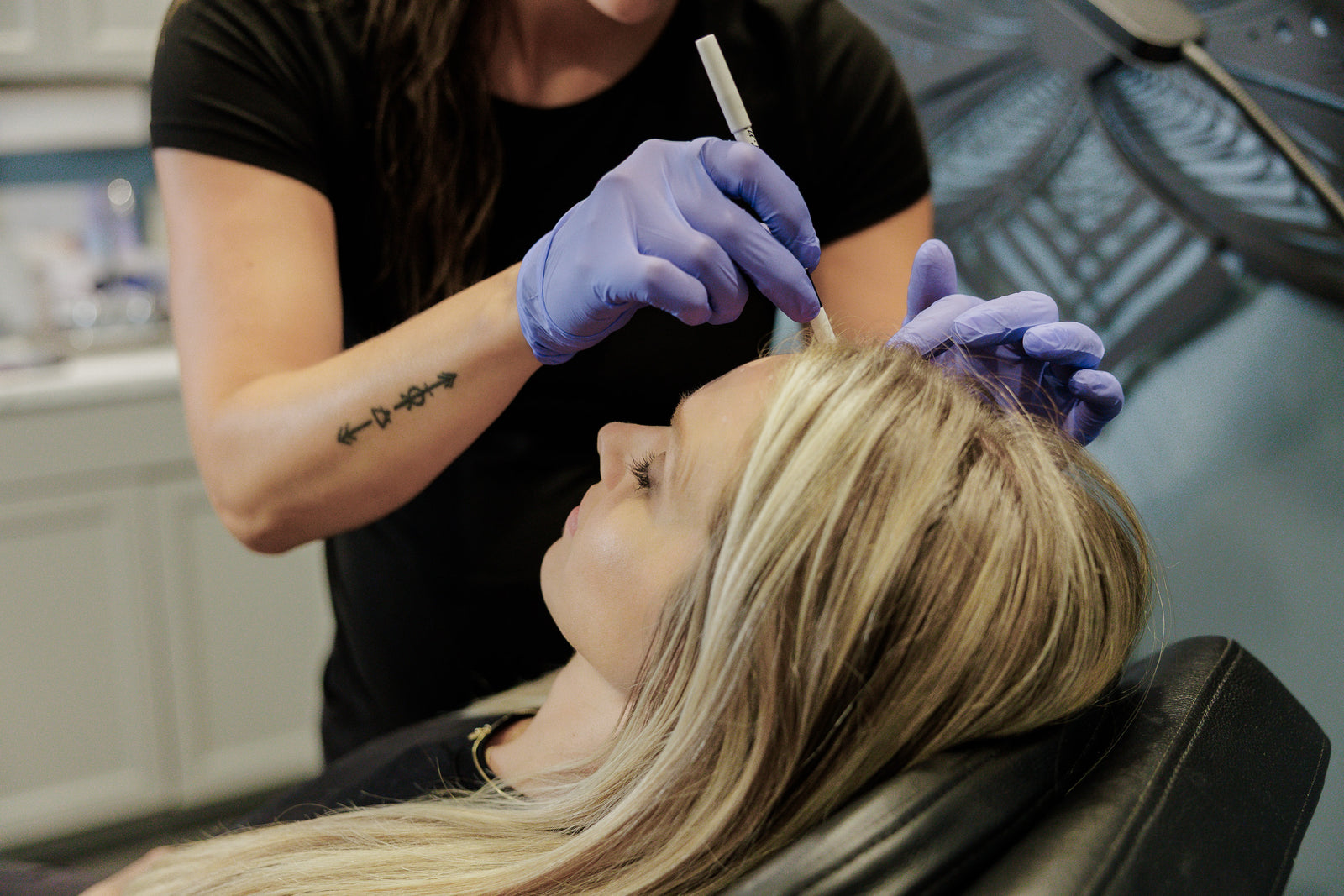 A woman with long blonde hair reclines in a salon chair while a stylist in a black shirt and purple gloves applies product to her hair. The salon interior is softly blurred in the background.