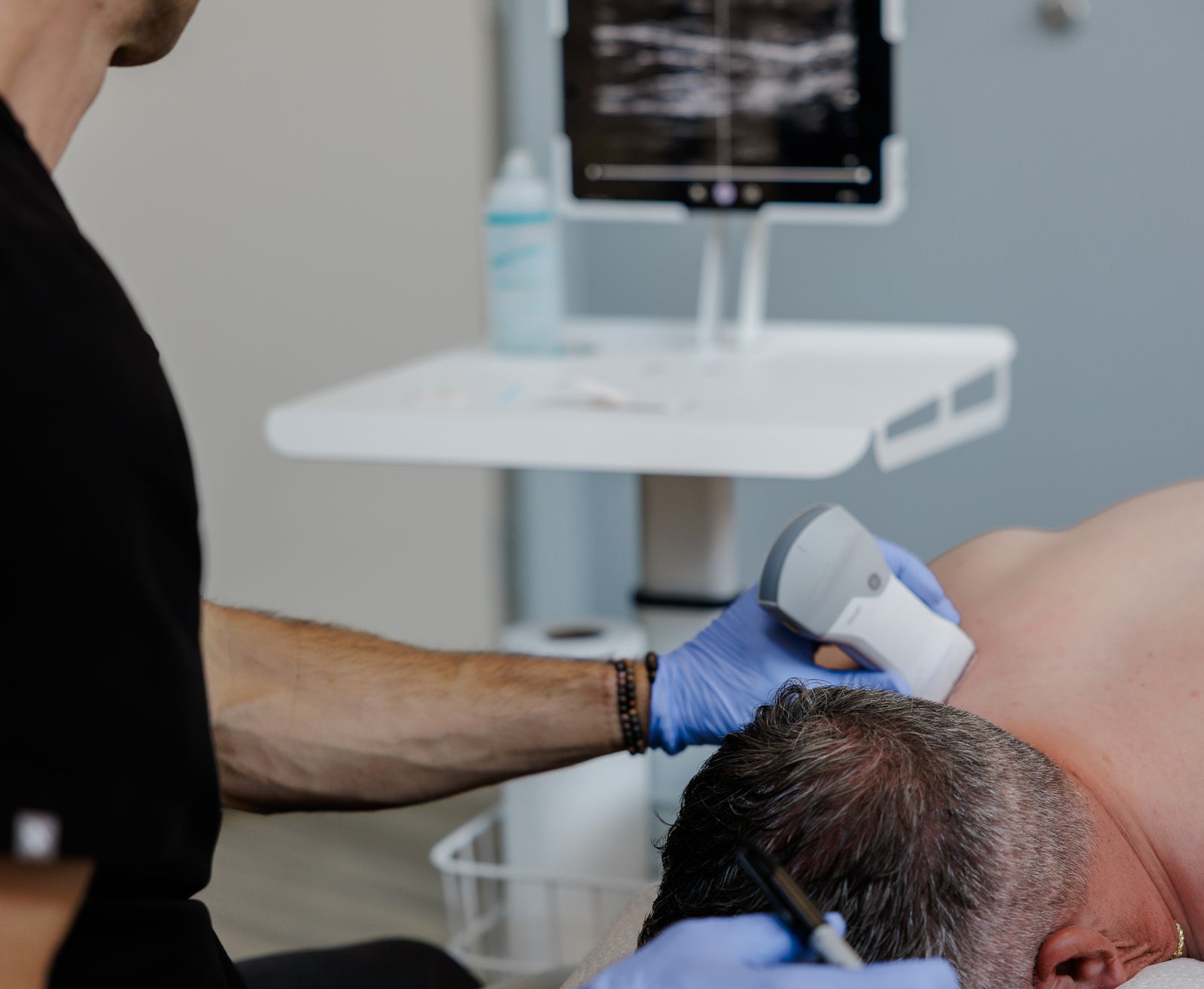 A healthcare provider wearing blue gloves uses a handheld ultrasound probe on a patient's upper back in a clinical room. A white ultrasound machine is visible in the background, along with a monitor and medical supplies.