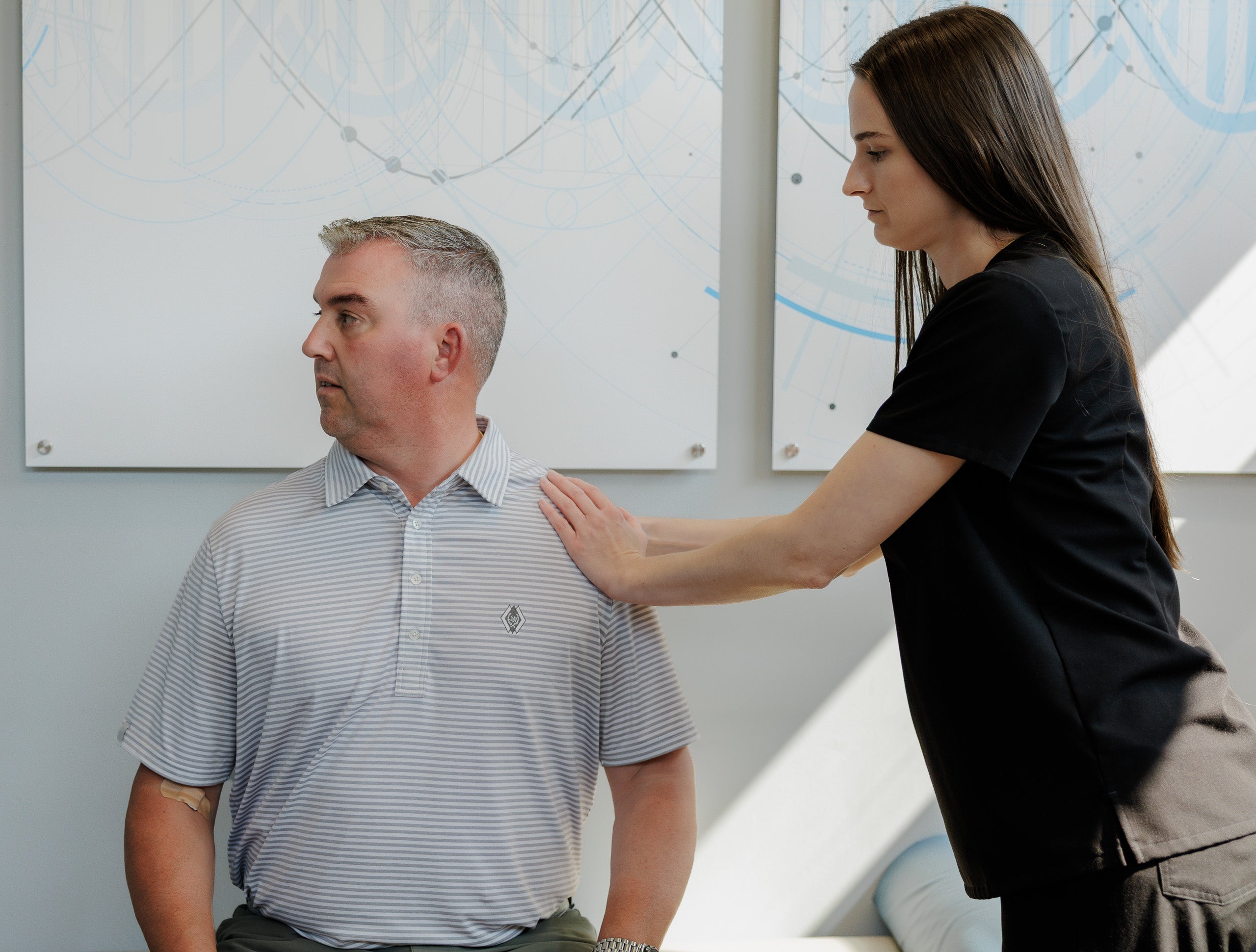 A woman in a black scrub top stands beside a seated man wearing a light blue and white striped polo shirt, placing her hand on his shoulder in what appears to be a clinical or office setting.