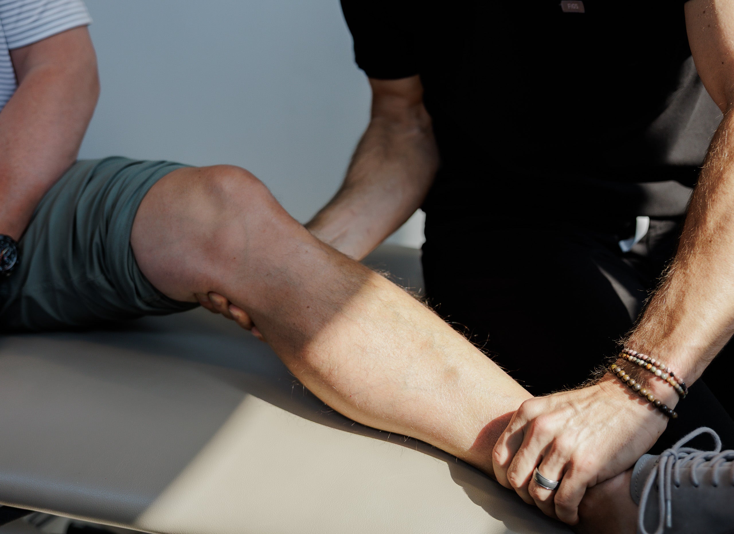 A close-up lifestyle shot of a physical therapist or doctor examining a person's leg and ankle, with the therapist's hands holding the ankle and the patient sitting on a treatment table.