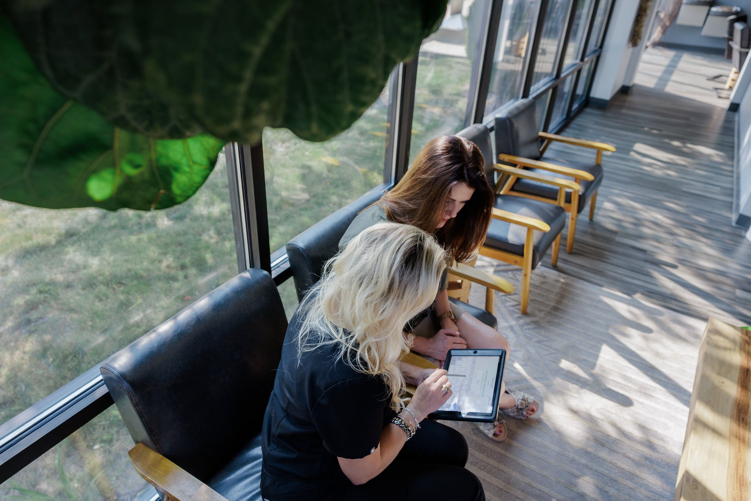 Two women sitting in a modern lounge area by large windows, looking at a tablet. The scene includes black leather chairs, light wood flooring, and a large green leaf in the foreground.