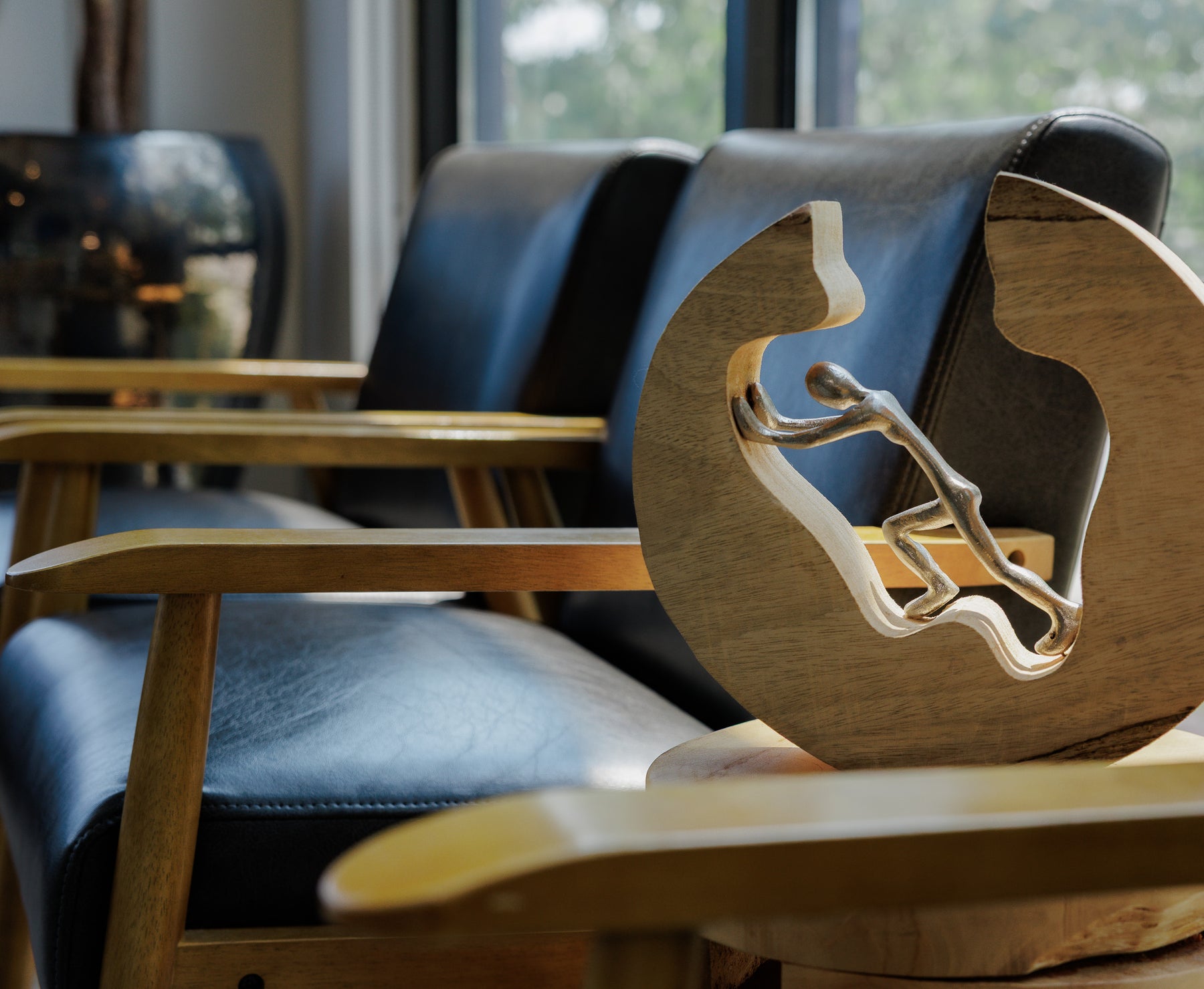 A close-up of a modern wooden chair with a sculptural circular backrest featuring a cutout of a seated figure, set in a real interior environment with other chairs and windows in the background.