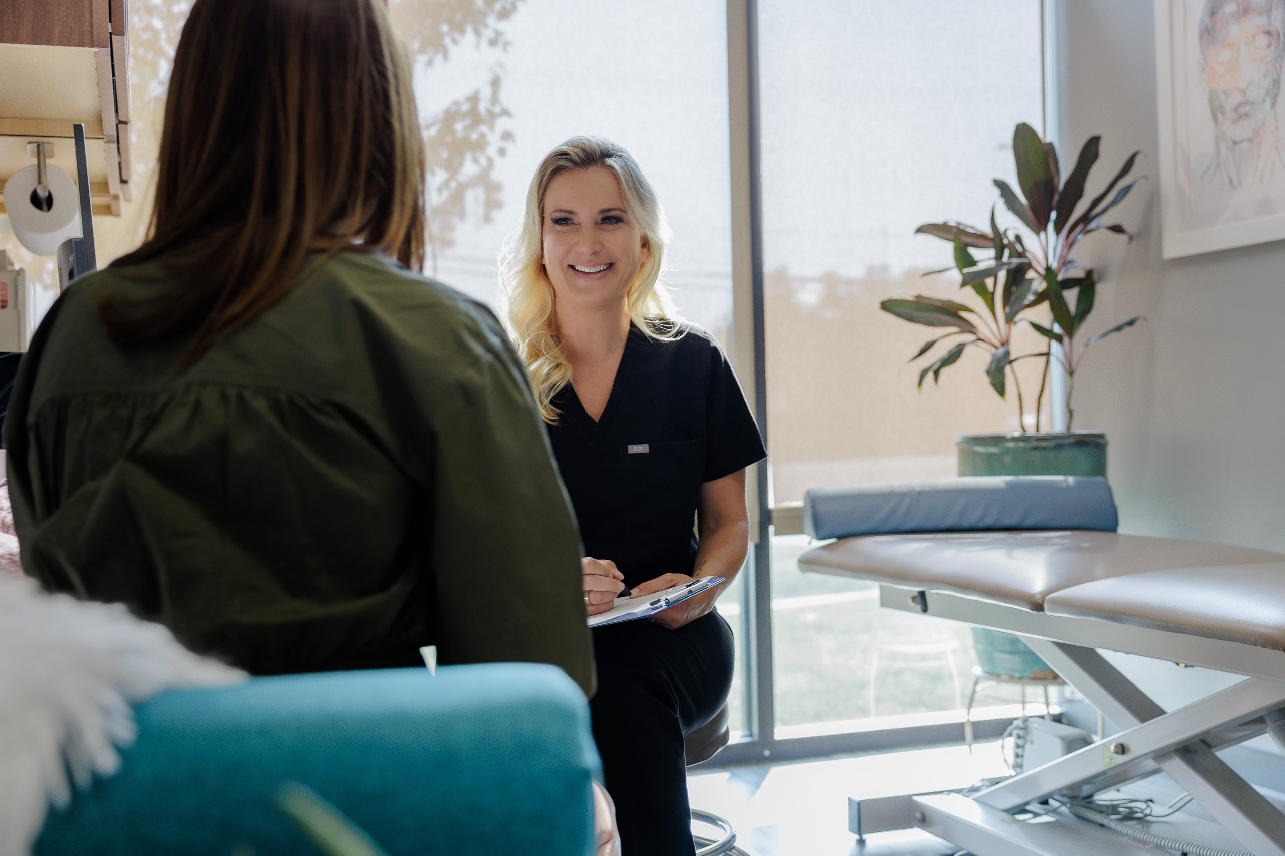 A smiling blonde woman in black medical scrubs sits in a bright clinic room, holding a clipboard and speaking with a client seated in the foreground. A treatment table and potted plant are visible in the background.