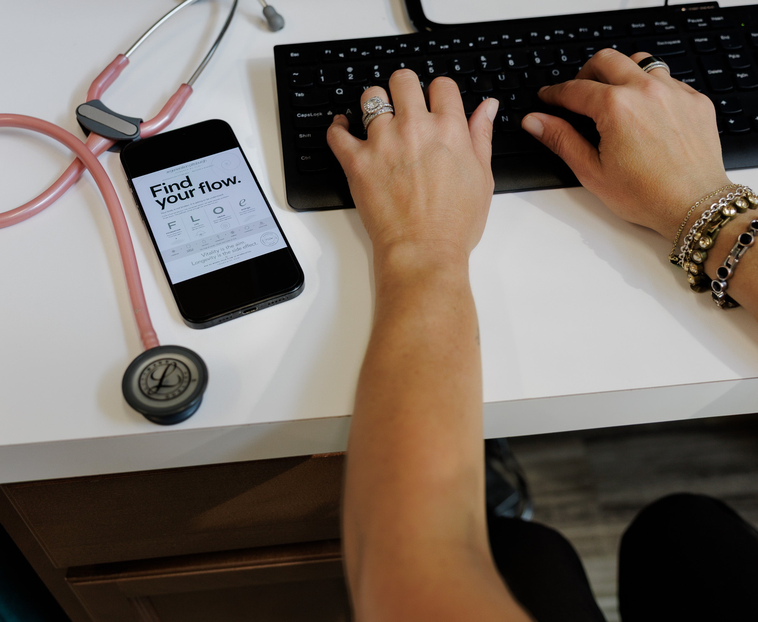A top-down view of a person's hands typing on a black computer keyboard on a white desk, with a pink stethoscope and a smartphone displaying 'Find your flow.' nearby.